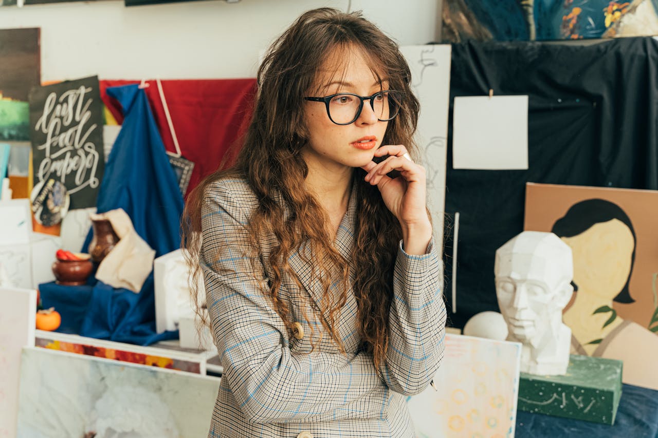 A thoughtful woman in an art studio surrounded by various paintings and sculptures.